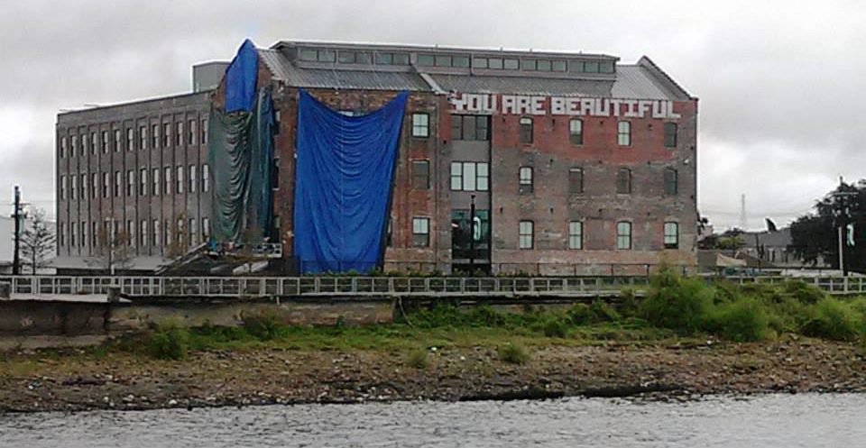 An old brick building, beaten by a storm. One corner is covered by an old blue tarp. The other side has block-letters that read, "You are beautiful".
